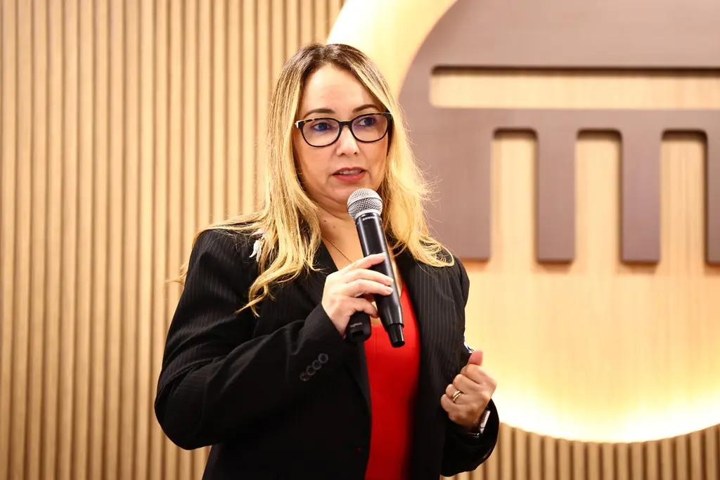 Foto horizontal em plano médio que mostra a juíza Ana Graziela Vaz em pé, falando ao microfone, no palco do auditório do Tribunal de Contas. Ela é uma mulher branca, de olhos castanhos, cabelos longos, lisos e loiros, usando blusa vermelha, blazer preto e óculos de grau.