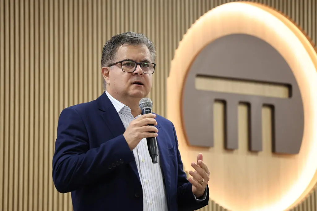 Foto horizontal em plano médio que mostra o coordenador de Planejamento do TJMT, Afonso Maciel, em pé, falando ao microfone, no auditório do Tribunal de Contas. Ele é um homem branco, de cabelos grisalhos, usando camisa branca, paletó azul marinho e óculos de grau.