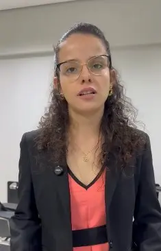 A imagem apresenta, em plano médio, uma mulher jovem de pele clara, cabelos escuros cacheados e óculos de grau, vestindo um blazer preto sobre uma blusa de tom coral