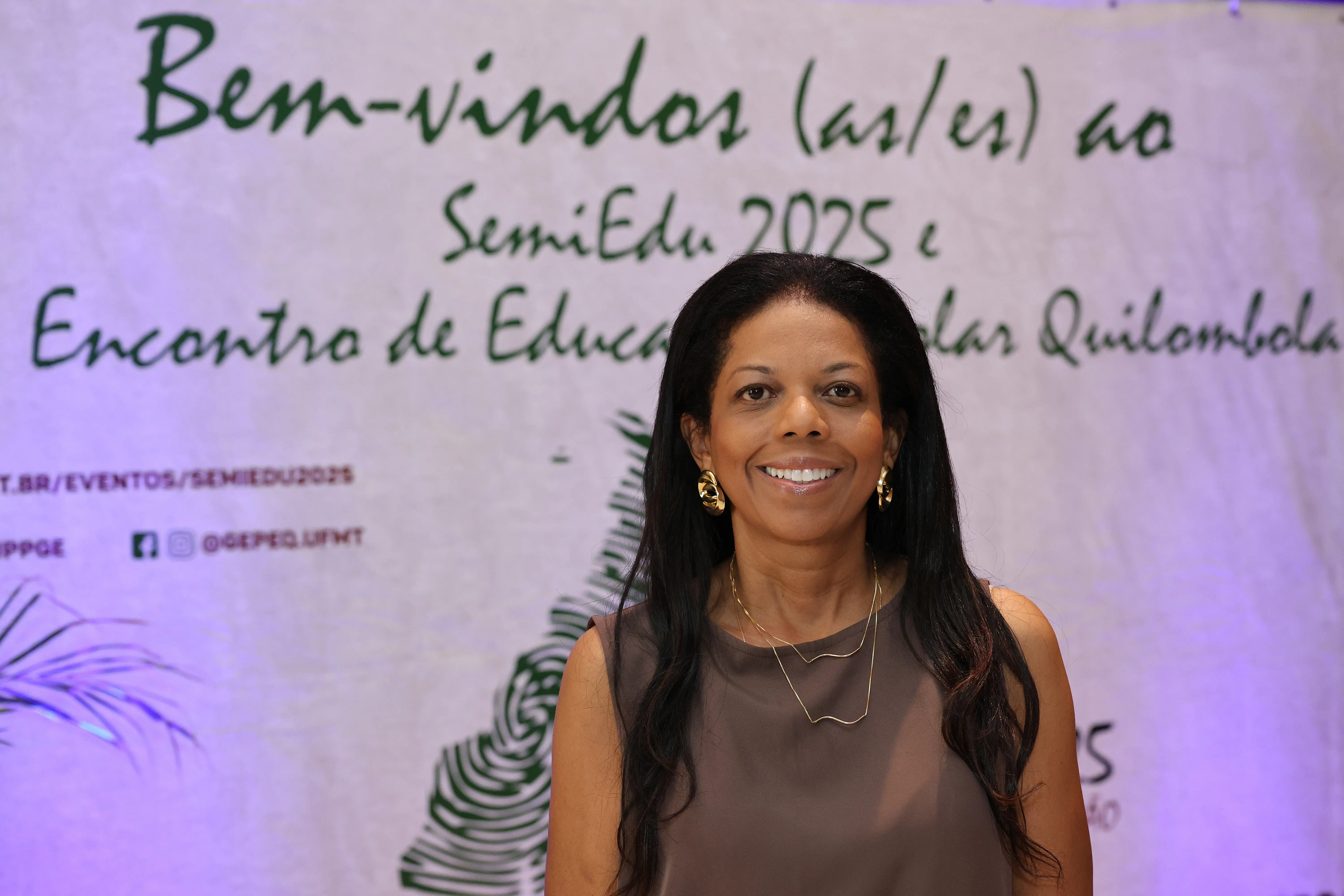 Dra. Renata Parreira. Retrato em close-up de uma mulher de cabelos longos e escuros, sorrindo. Ela usa brincos grandes e correntes finas. Ao fundo, um banner do 