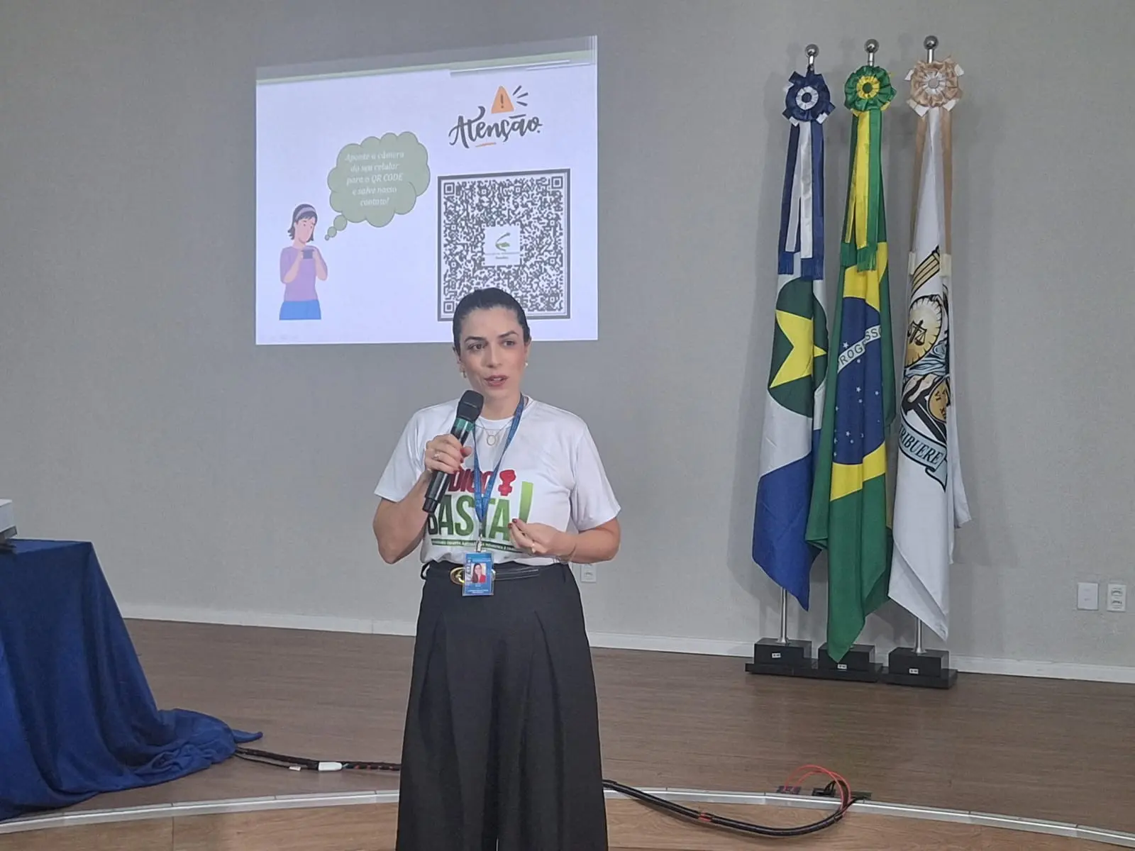 Foto horizontal em plano aberto que mostra a psicóloga do Núcleo Thays Machado, Luciana Edeliz, durante palestra no auditório do fórum de Cuiabá. Ela é uma mulher branca, magra, de olhos e cabelos escuros, cabelos presos, usando camiseta da campanha Eu Digo Basta e calça preta.