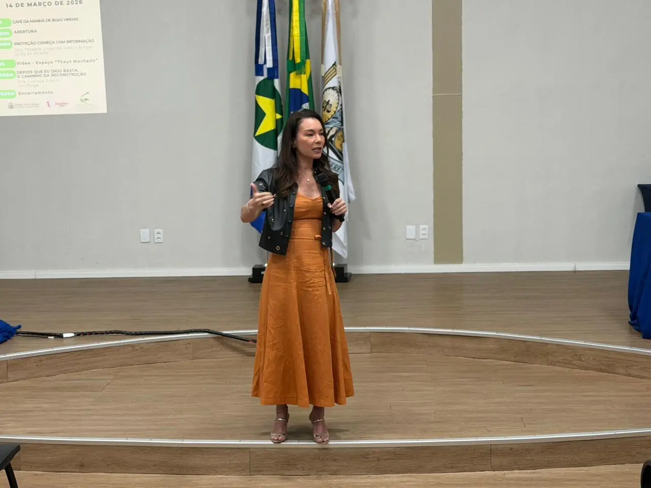 Foto horizontal em plano aberto que mostra a juíza Hanae Yamamura falando ao microfone, em pé, no auditório do fórum de Cuiabá. Ela é uma mulher de pele clara, magra, cabelos castanhos, lisos e longos, com traços orientais, usando vestido marrom e camisa preta de couro e manga curta