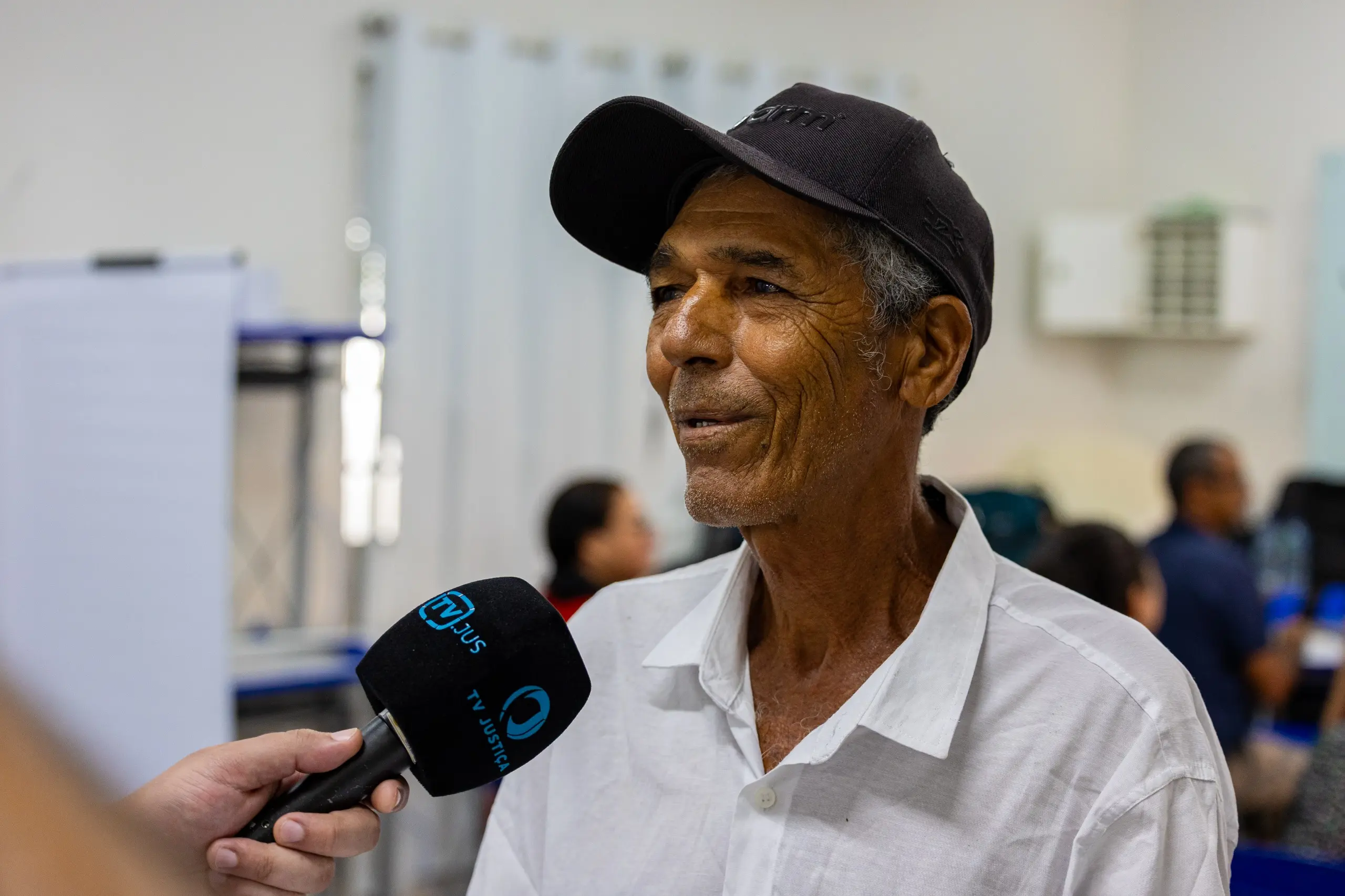 Homem pardo, de perfil, com boné preto e camisa branca social, sorri enquanto fala ao microfone da TV Justiça. Ao fundo, um ambiente de atendimento com outras pessoas e móveis de escritório.