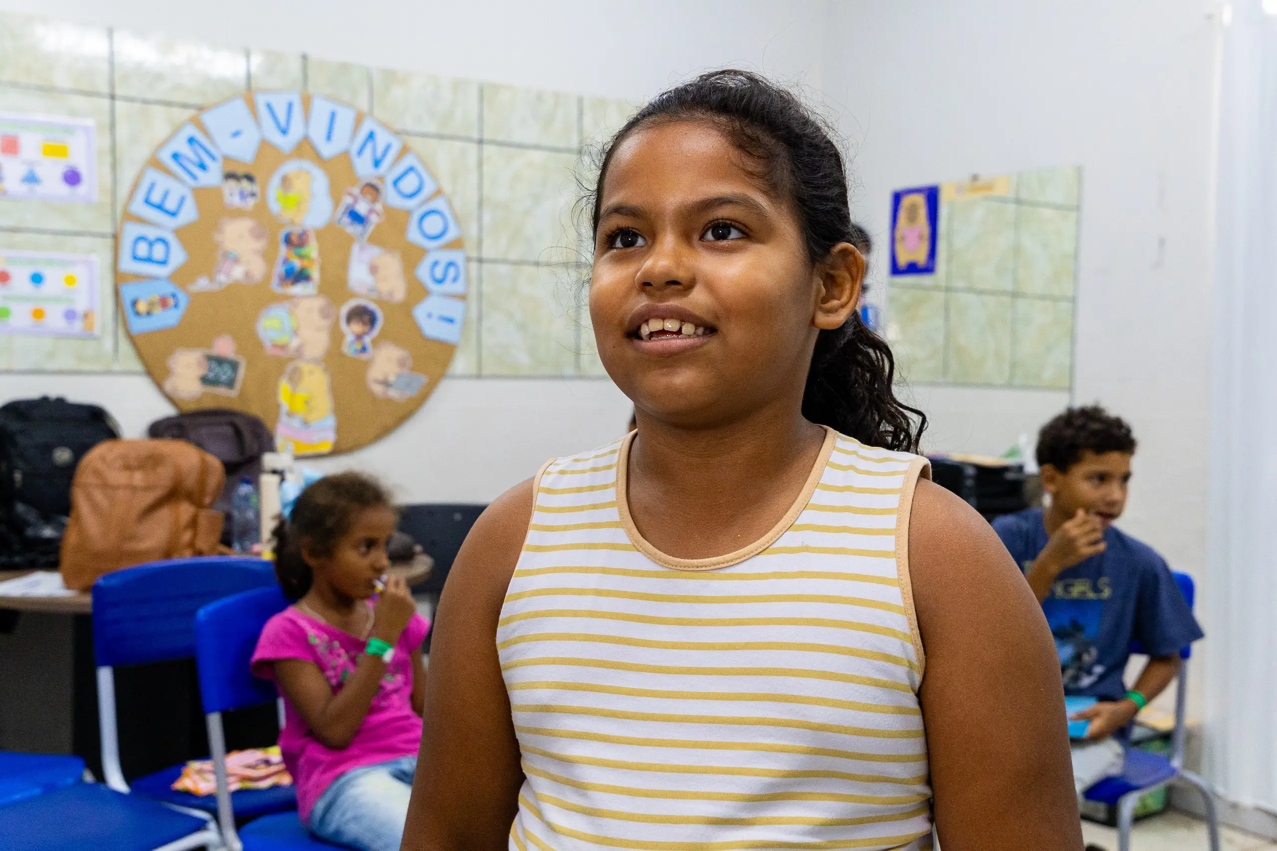 Menina em primeiro plano, sorridente, veste regata listrada de amarelo e branco. Ao fundo, outras crianças sentadas em cadeiras azuis e um painel circular escrito 