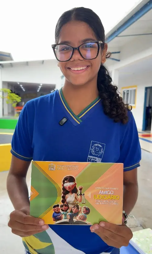 Foto vertical que mostra a aluna Isabella Vitória, de 12 anos, da cintura pra cima, no pátio da escola, sorrindo e mostrando a cartilha do Nosso Judiciário. Ela é uma menina negra, de cabelos castanhos e cacheados, usando uniforme azul da escola e óculos de grau. 