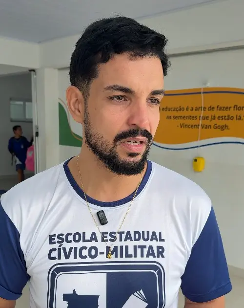 Foto vertical que mostra o coordenador pedagógico da Escola Estadual Ana Maria do Couto, Osvaldo Ribeiro de Souza Neto, no pátio da unidade. Ele é um homem jovem, branco, de olhos, cabelo e barba pretos, usando camiseta da escola. 