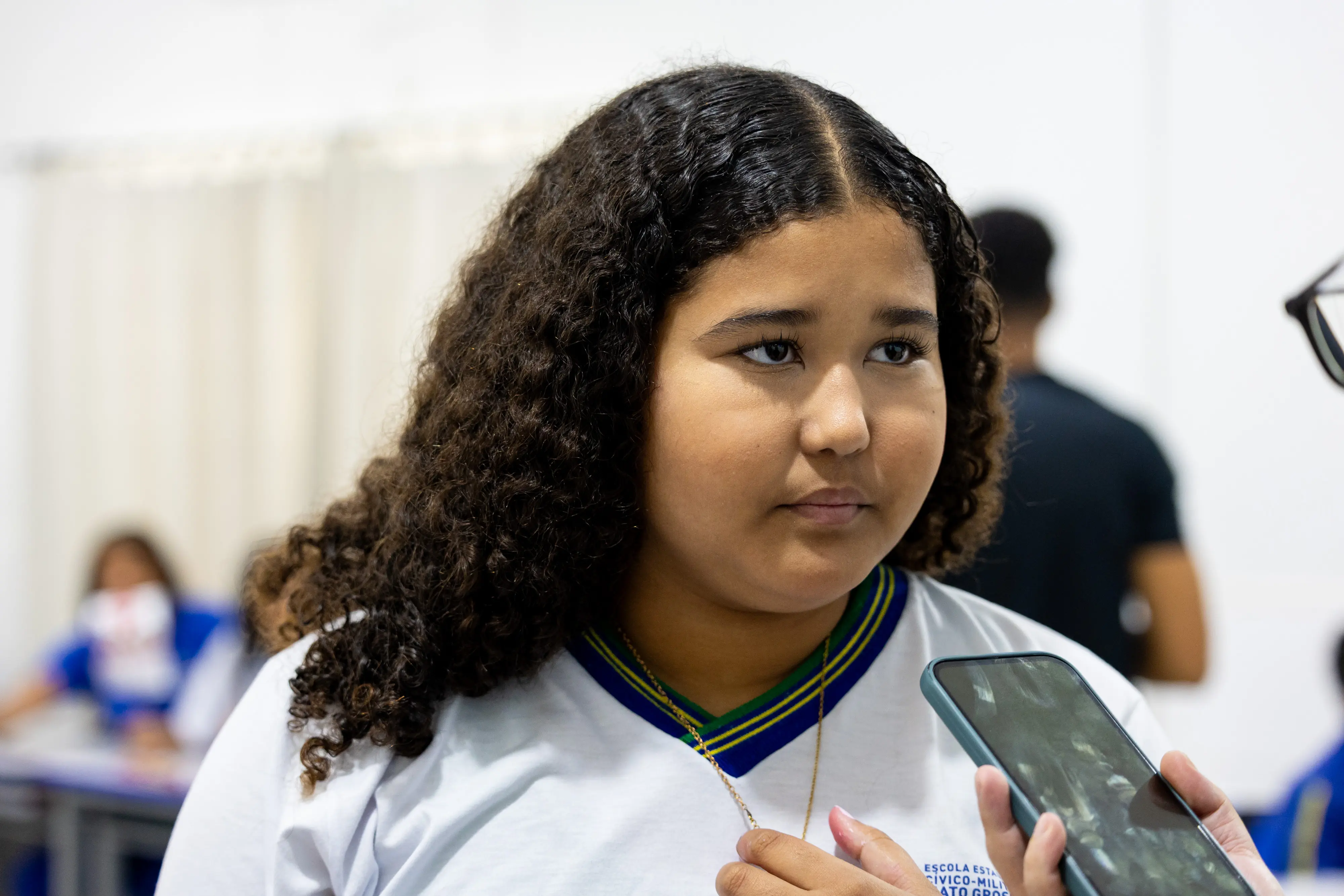 Close de uma jovem de cabelos cacheados e escuros, vestindo uniforme escolar branco com detalhes em azul e verde. Ela olha para o lado enquanto uma pessoa segura um celular próximo, como em uma entrevista.