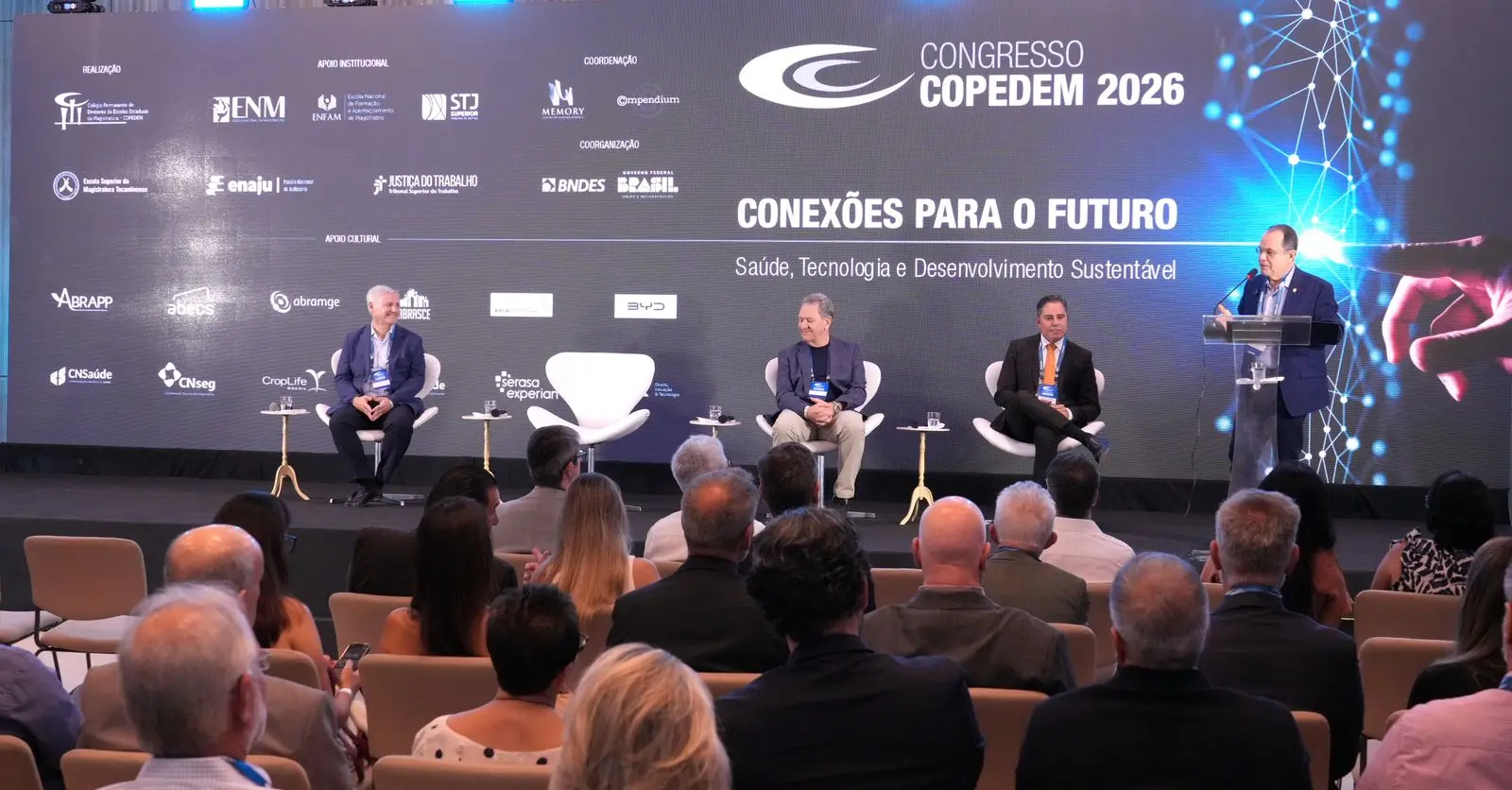 Foto colorida. Homem fala ao microfone, da tribuna. Ele veste terno escuro e camisa branca. Ao lado dele, três outros homens sentam em cadeiras alinhadas uma ao lado da outra