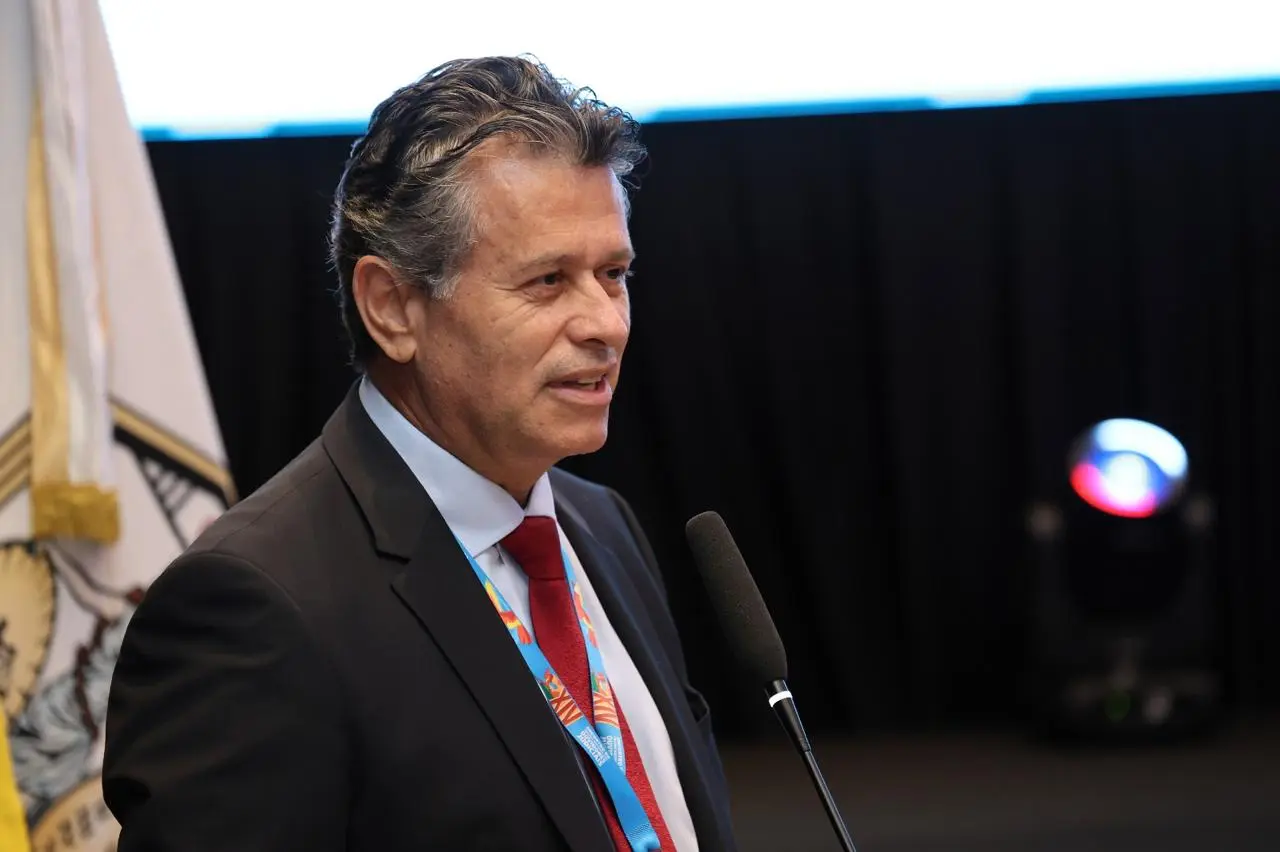 Homem de terno escuro e gravata vermelha discursa ao microfone, em close. Usa crachá do evento e demonstra expressão séria e concentrada, com fundo escuro e iluminação destacando o orador.