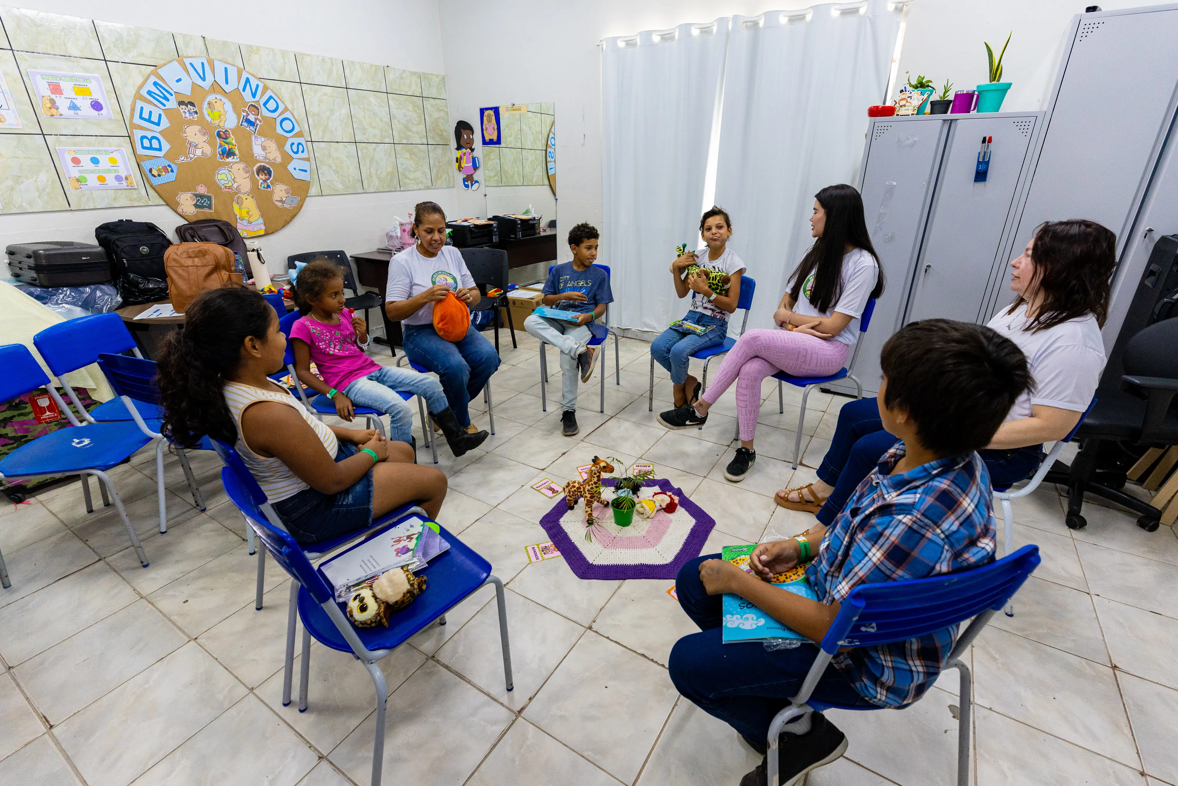 Crianças e adultos formam uma roda em cadeiras azuis em uma sala. No centro do círculo, há um tapete com brinquedos. As paredes têm cartazes infantis e o clima é de conversa e atividade lúdica.