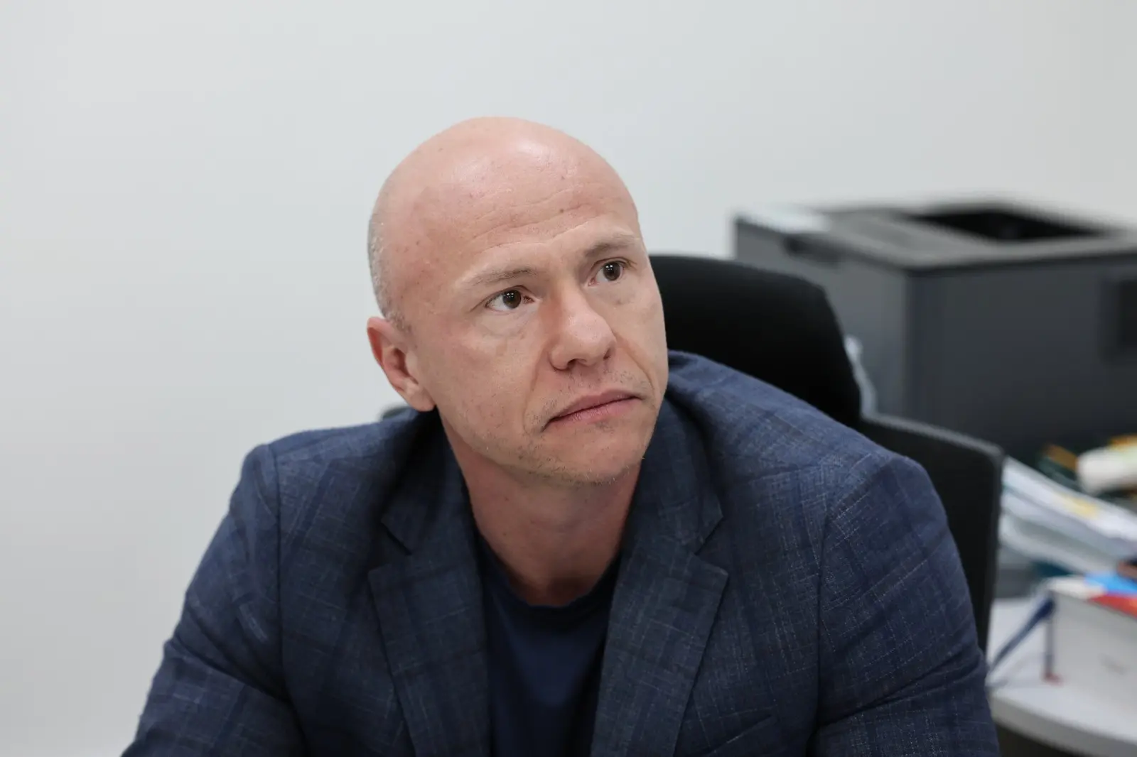 Homem careca, de blazer azul, está sentado em mesa de escritório. Ele olha levemente para o lado, com expressão atenta. Ao fundo, equipamentos de trabalho indicam ambiente profissional.