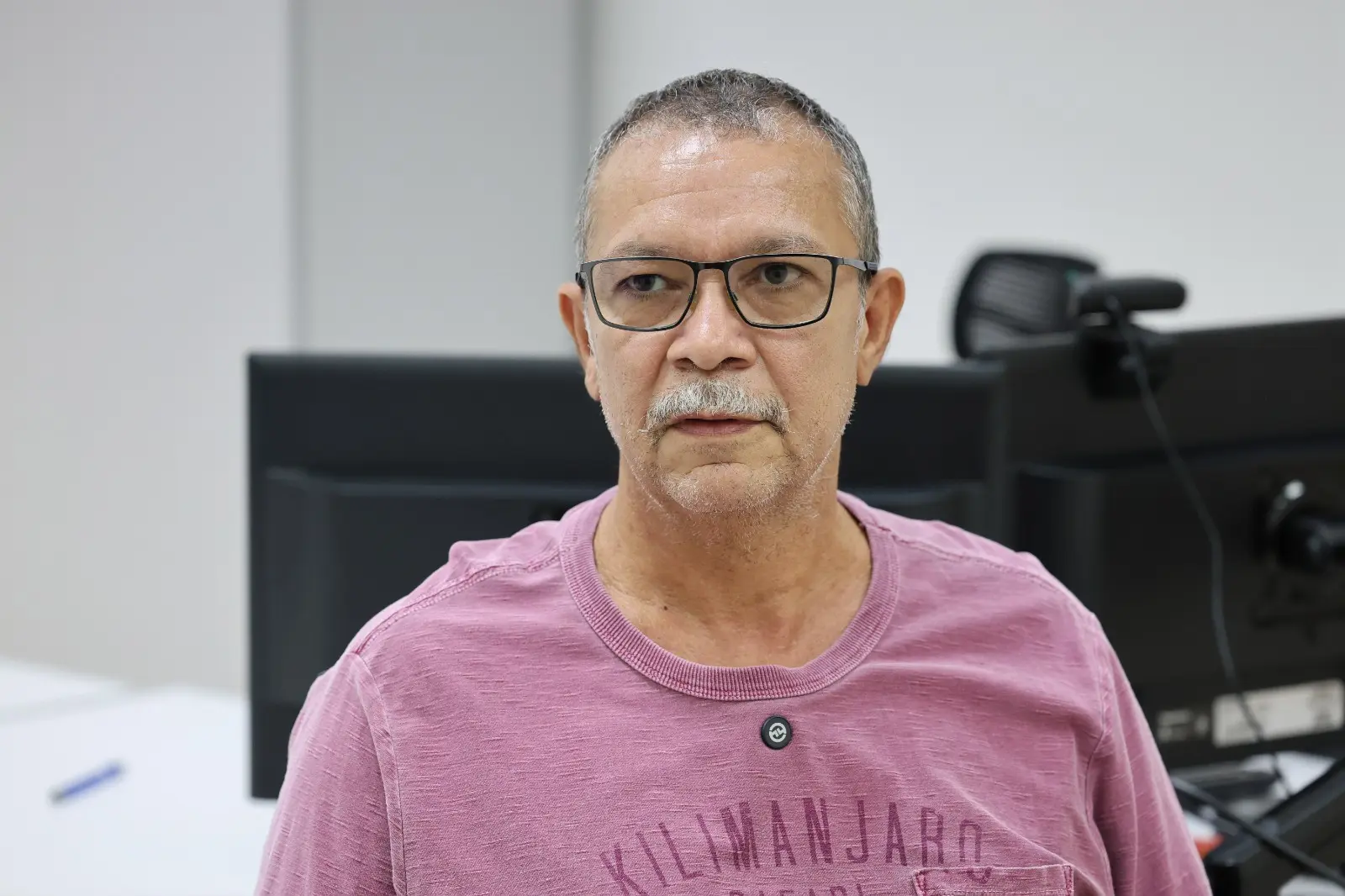 Homem de cabelos grisalhos e óculos aparece em primeiro plano, usando camiseta rosa. Ao fundo, há computadores e cadeiras, indicando ambiente de trabalho. Sua expressão é neutra e direta.