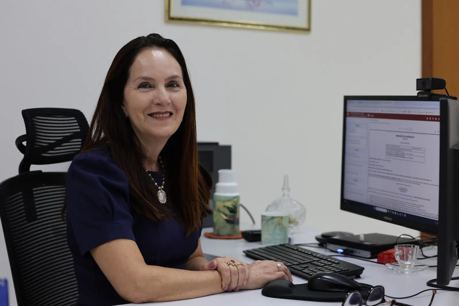 Mulher de cabelos escuros e longos, veste blusa azul e sorri para a câmera. Está sentada em escritório, com computador e monitor ao lado. O ambiente é claro, organizado e com itens de uso profissional.