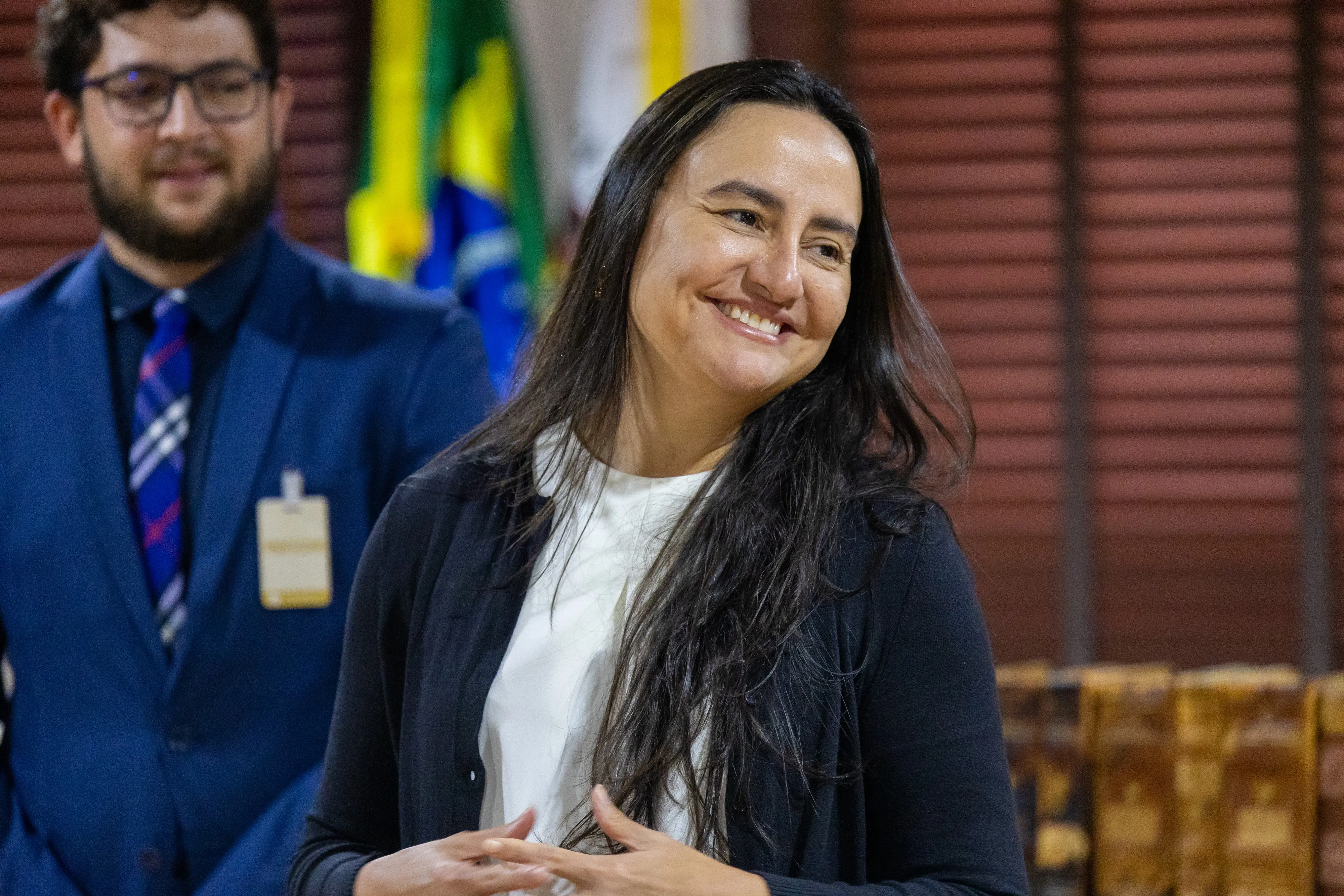 Uma mulher de cabelos longos e escuros, veste blusa branca e cardigan azul-marinho, sorri amplamente. Ao fundo, um jovem de terno azul e a bandeira do Brasil.