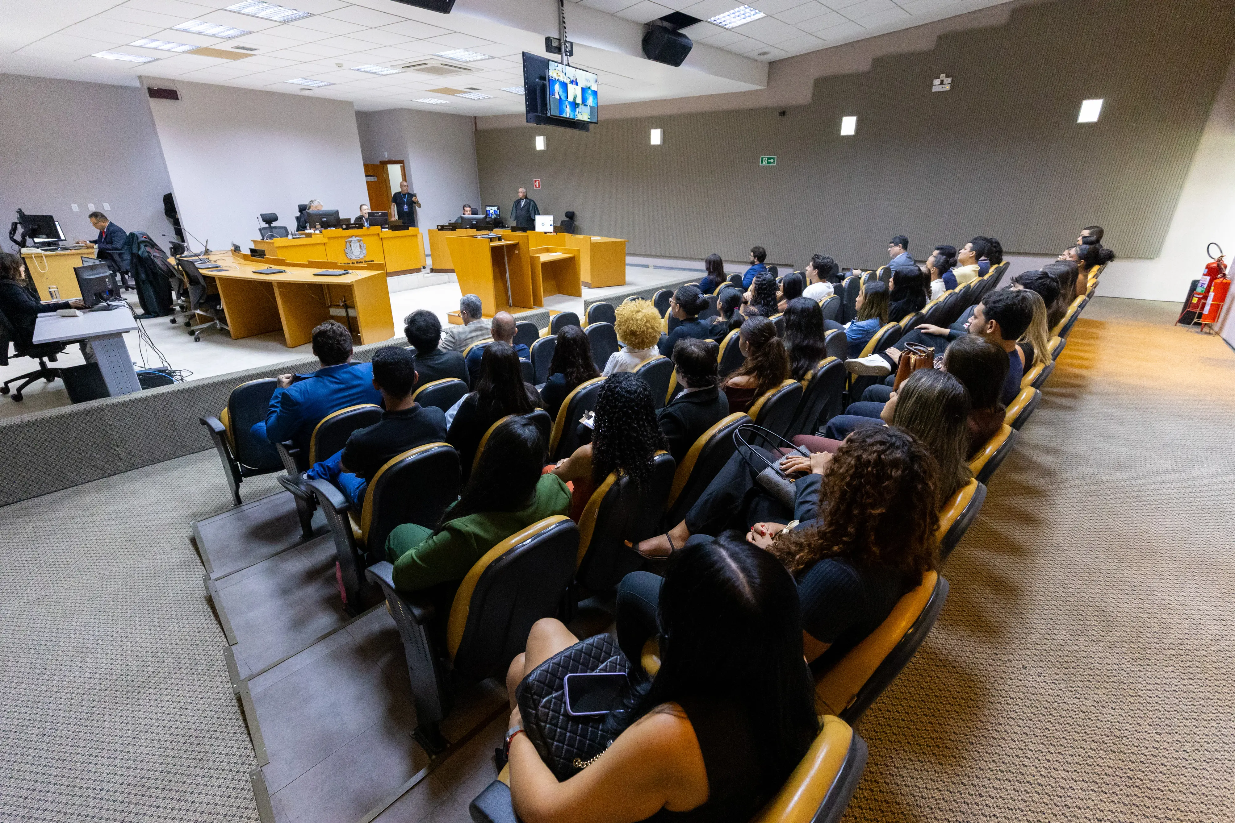 Dezenas de estudantes assistem a uma sessão do Tribunal, sentados em cadeiras amarelas, enquanto magistrados trabalham ao fundo sobre uma bancada elevada.