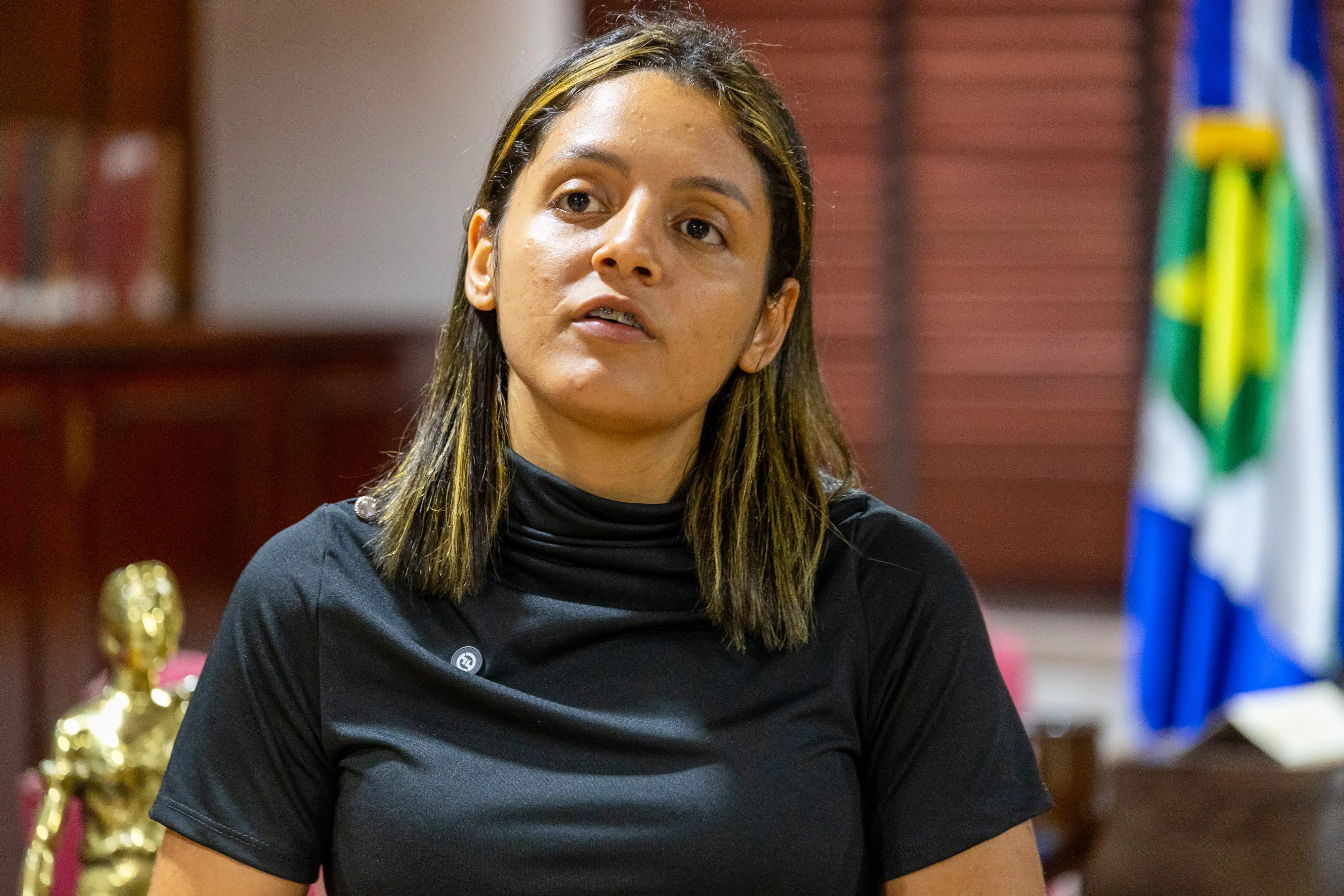 Uma mulher de cabelos escuros com mechas loiras, veste blusa preta de gola alta e observa algo à sua frente. Ao fundo, estantes com livros e a bandeira de Mato Grosso.