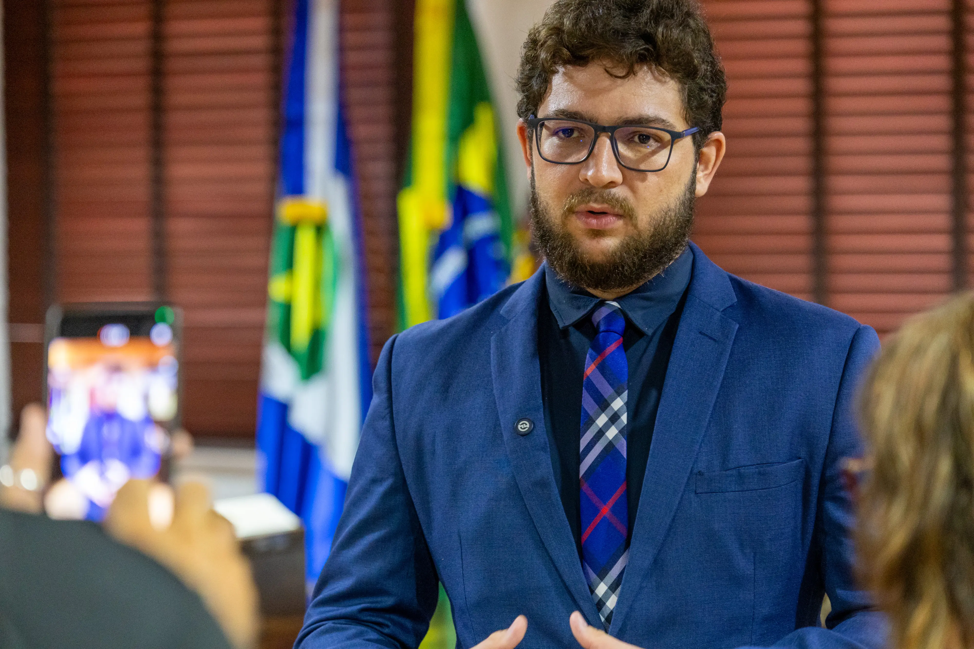 Um homem de óculos, barba e cabelos cacheados, veste terno azul e gravata xadrez azul e vermelha, fala gesticulando com as mãos. Ao fundo, as bandeiras do Brasil e de Mato Grosso.