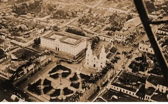 Vista aérea antiga do centro de Cuiabá, com praça arborizada, igreja ao centro e prédios baixos ao redor. Ruas organizadas e poucos veículos mostram a cidade em formação.