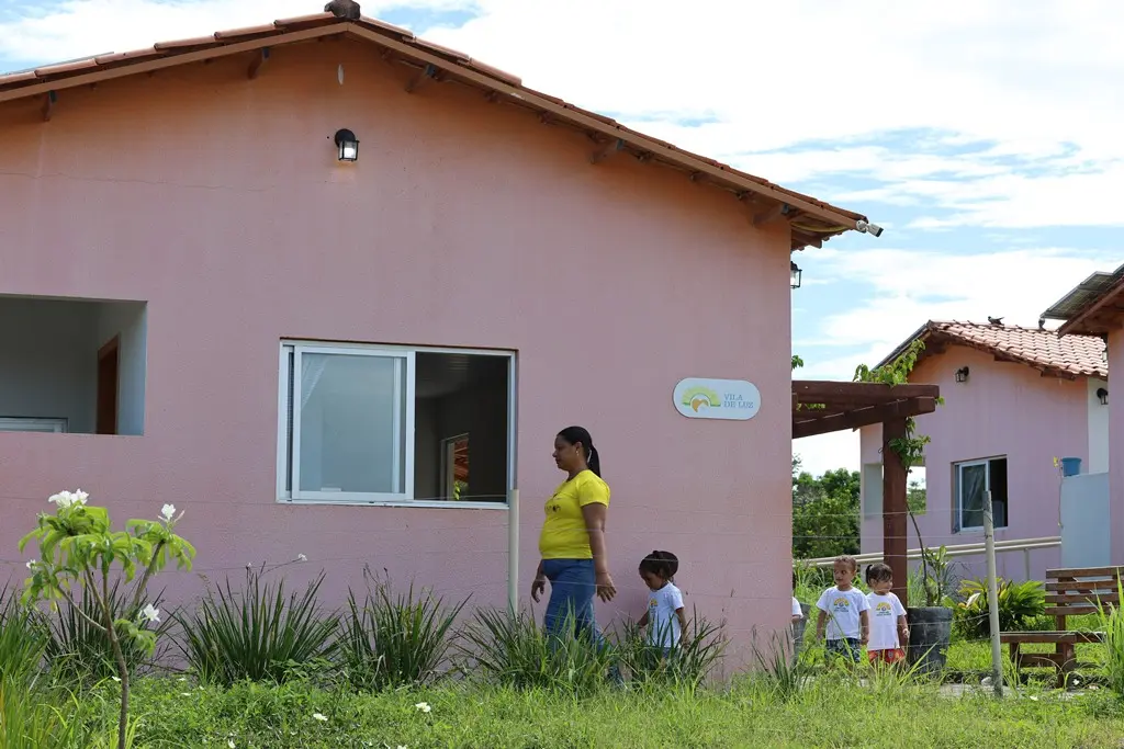 Foto horizontal colorida que mostra em primeiro plano uma casa da Vila de Luz, com outra casa ao fundo. Uma professora e um grupo de alunos andam em fila indiana em frente à casa, que é rodeada por grama e plantas.