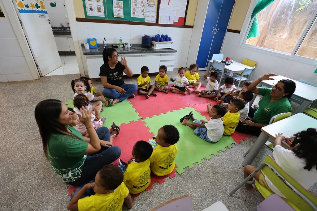 Foto horizontal colorida que mostra uma sala de aula da creche Nina Zaque com várias crianças e três professoras sentadas em círculo, sobre tatames coloridos no chão.