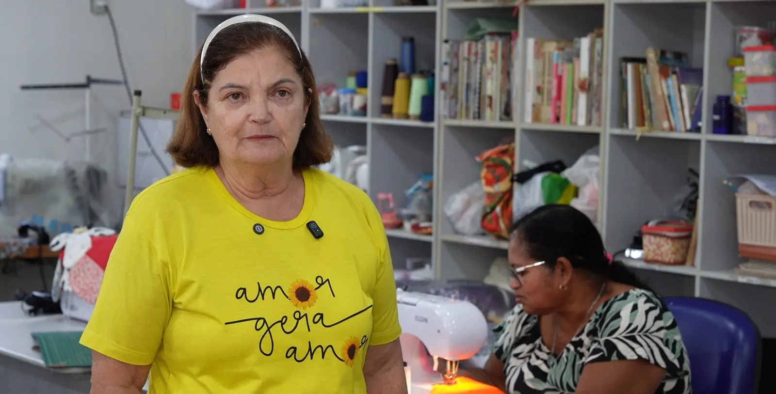 Foto horizontal que mostra a presidente das Obras Sociais Seara de Luz, Elione Fátima, em uma oficina de costura. Ela é uma mulher branca, se cabelos castanhos na altura dos ombros, usando camiseta amarela com a frase 