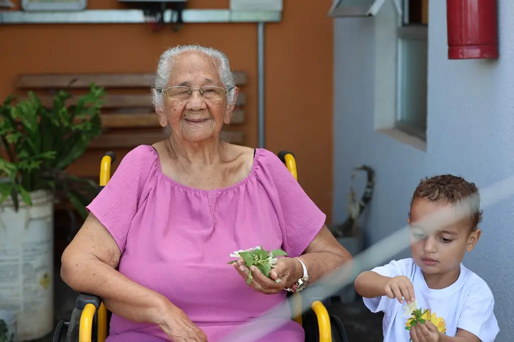 Foto horizontal colorida que mostra a idosa Maria sentada em uma cadeira de rodas, sorrindo para a foto e segurando flores. Ela usa um vestido rosa. Ao lado dela há um menininho de 3 anos de idade, segurando flores.