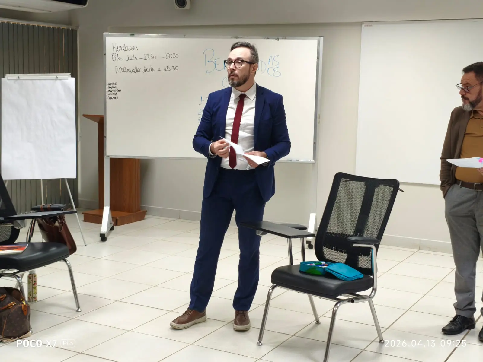 Um homem de pele clara, cabelos curtos escuros e barba grisalha sorri para a câmera vestindo um blazer marrom sobre uma camisa polo mostarda. Ele está posicionado em um corredor moderno com guarda-corpo branco