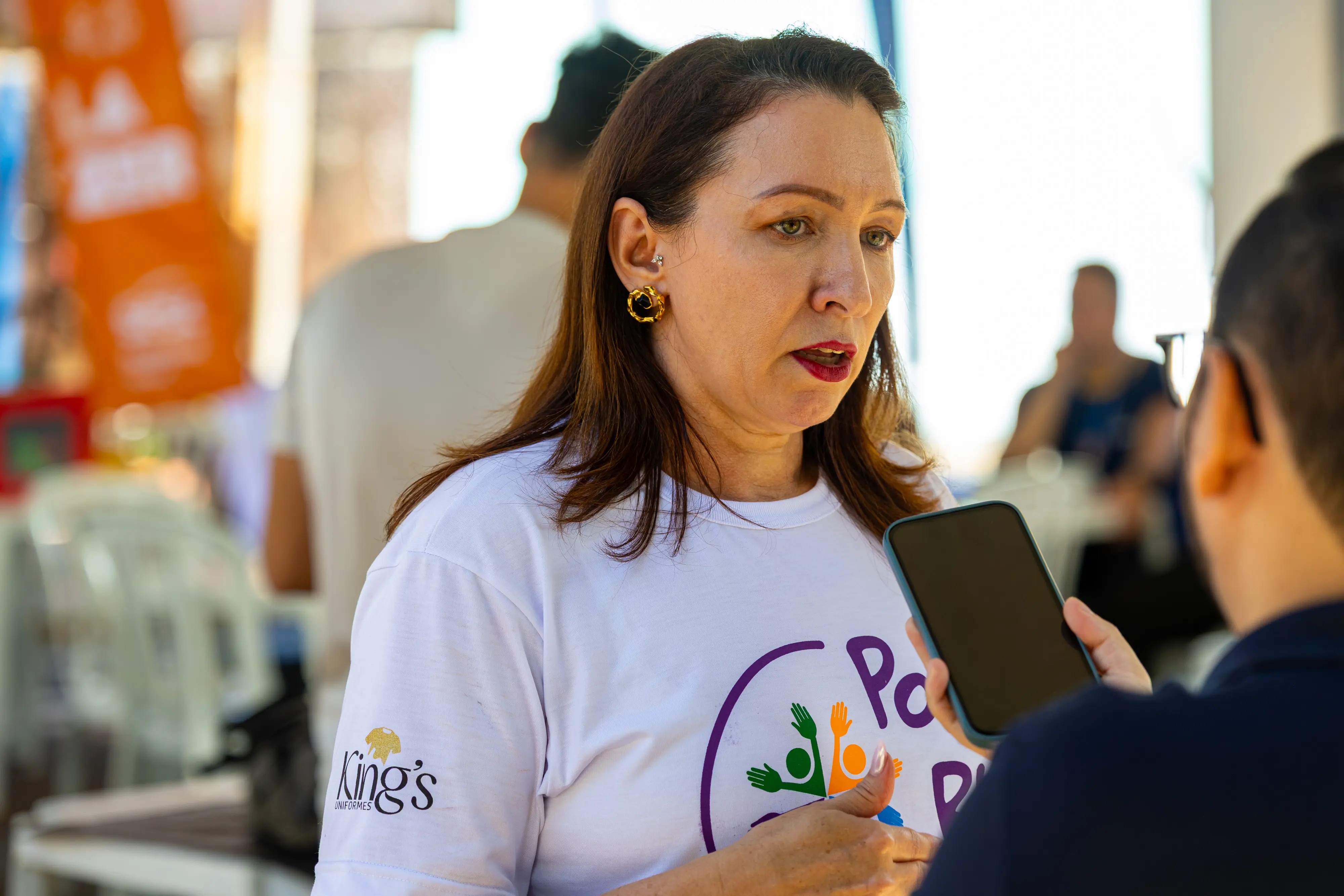 Uma mulher de cabelos castanhos e camiseta branca com logotipo colorido, fala durante uma entrevista. Um repórter de óculos segura um celular próximo a ela para gravar o áudio.