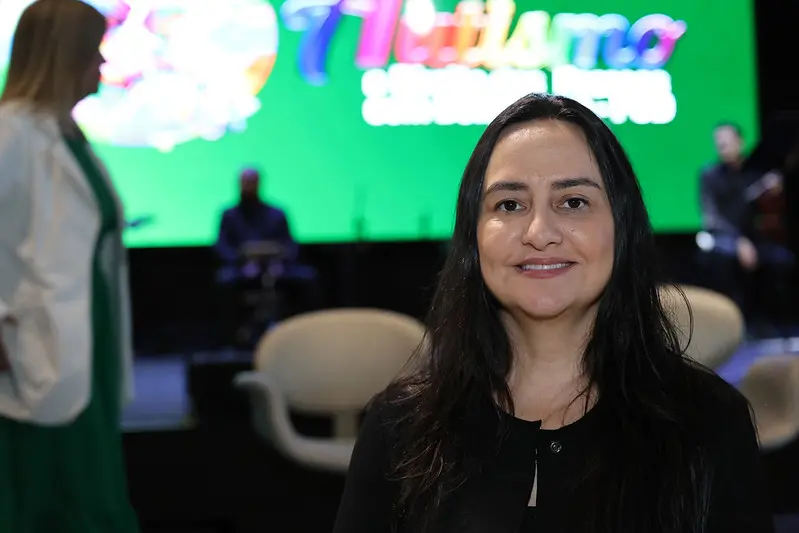 Mulher de cabelos longos posa sorrindo em primeiro plano. Ao fundo, palco com cadeiras e telão verde do evento TJMT Inclusivo, com pessoas desfocadas ao redor.