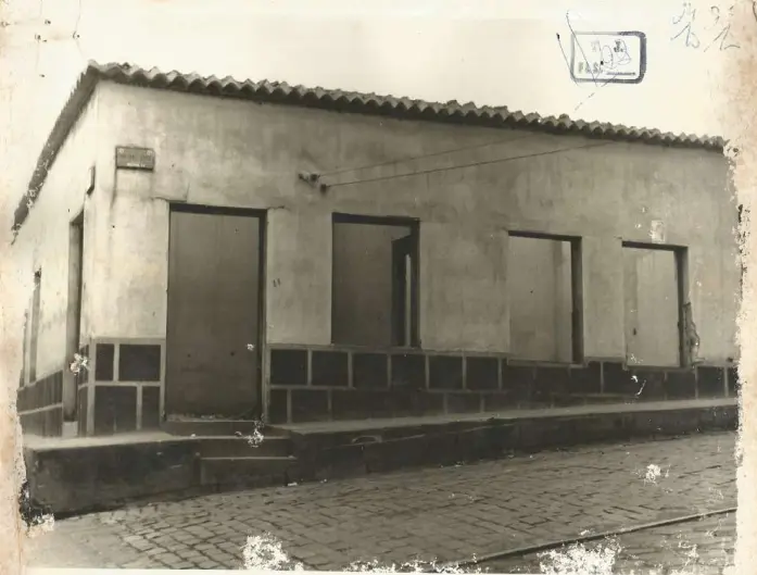 Foto antiga em preto e branco de uma casa de esquina térrea. Possui telhado simples, uma porta com degraus e três janelas retangulares sem vidros. A rua à frente é de paralelepípedos.