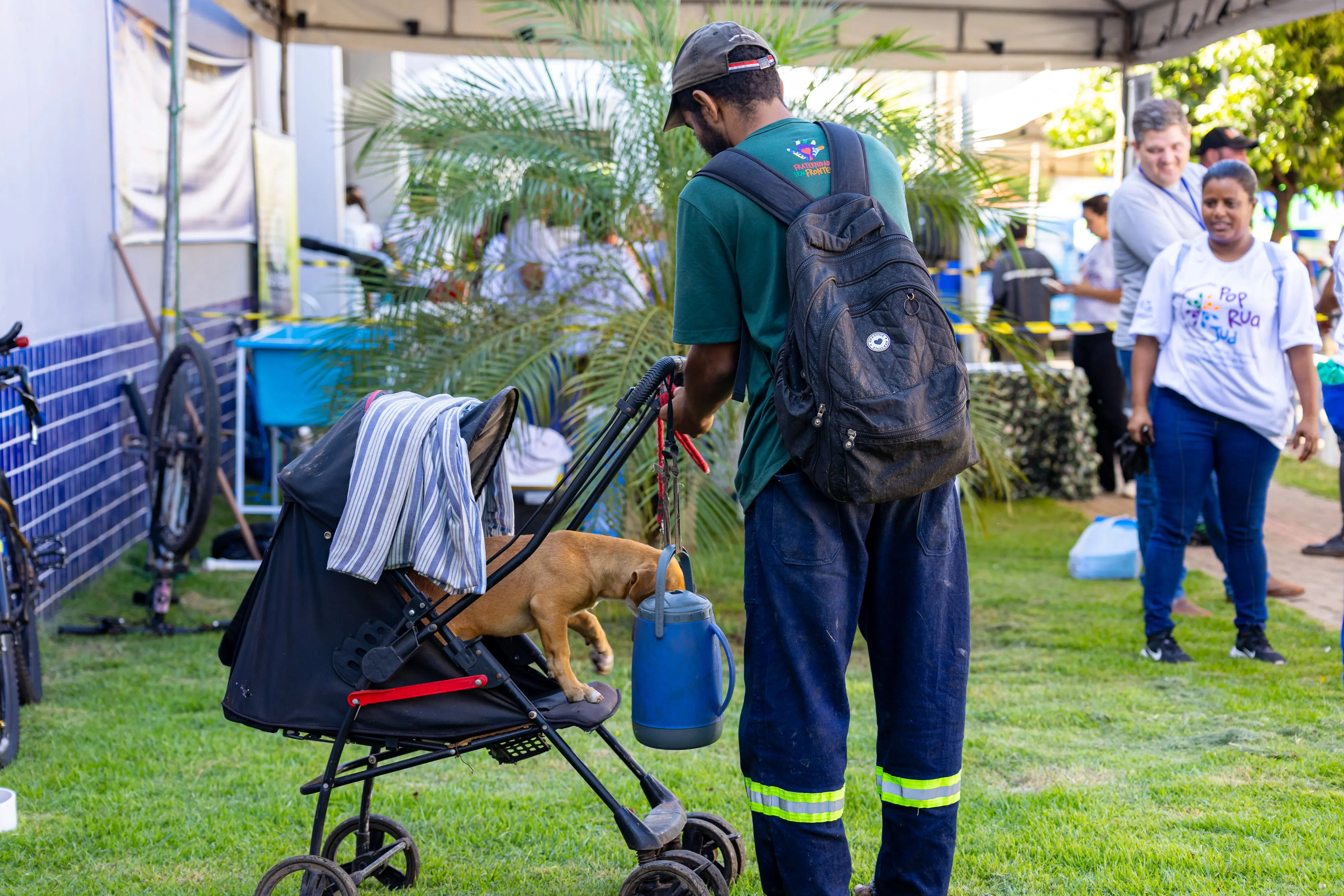 Um homem de costas, vestindo camiseta verde e mochila, empurra um carrinho de bebê preto onde está um cachorro caramelo. Ele segura uma garrafa térmica azul. O cenário é um gramado externo.