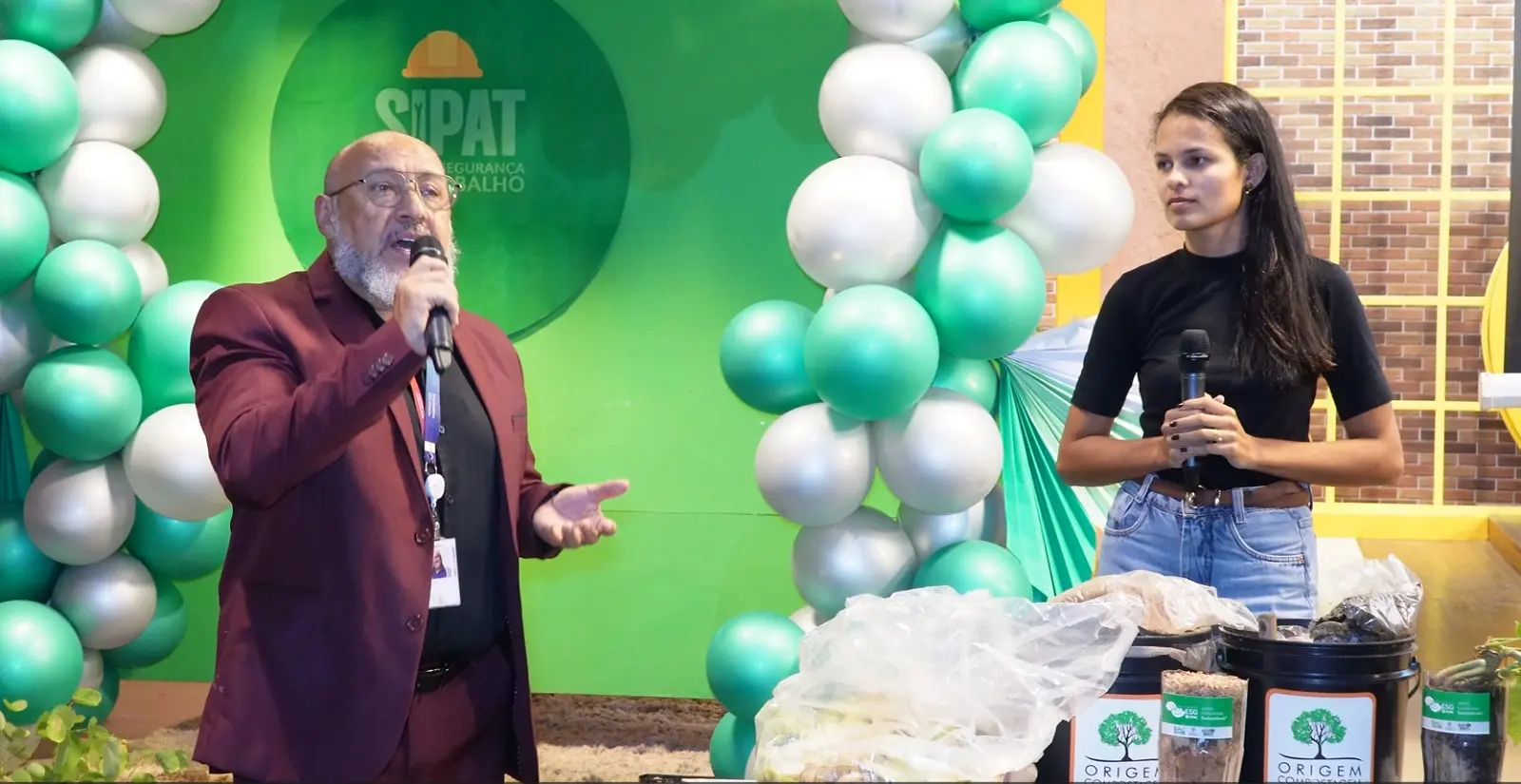 Homem de terno vinho fala ao microfone em um palco decorado com balões verdes e brancos. Ao lado, uma jovem observa. À frente, baldes com insumos.
