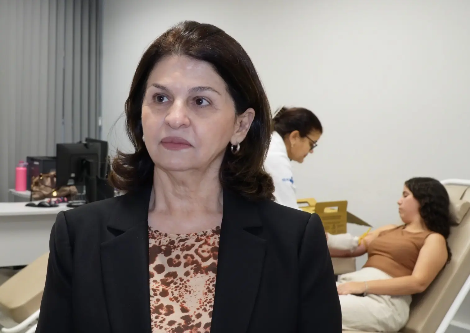 Mulher em primeiro plano. Ao fundo, uma jovem sentada em uma cadeira hospitalar realiza a doação de sangue.