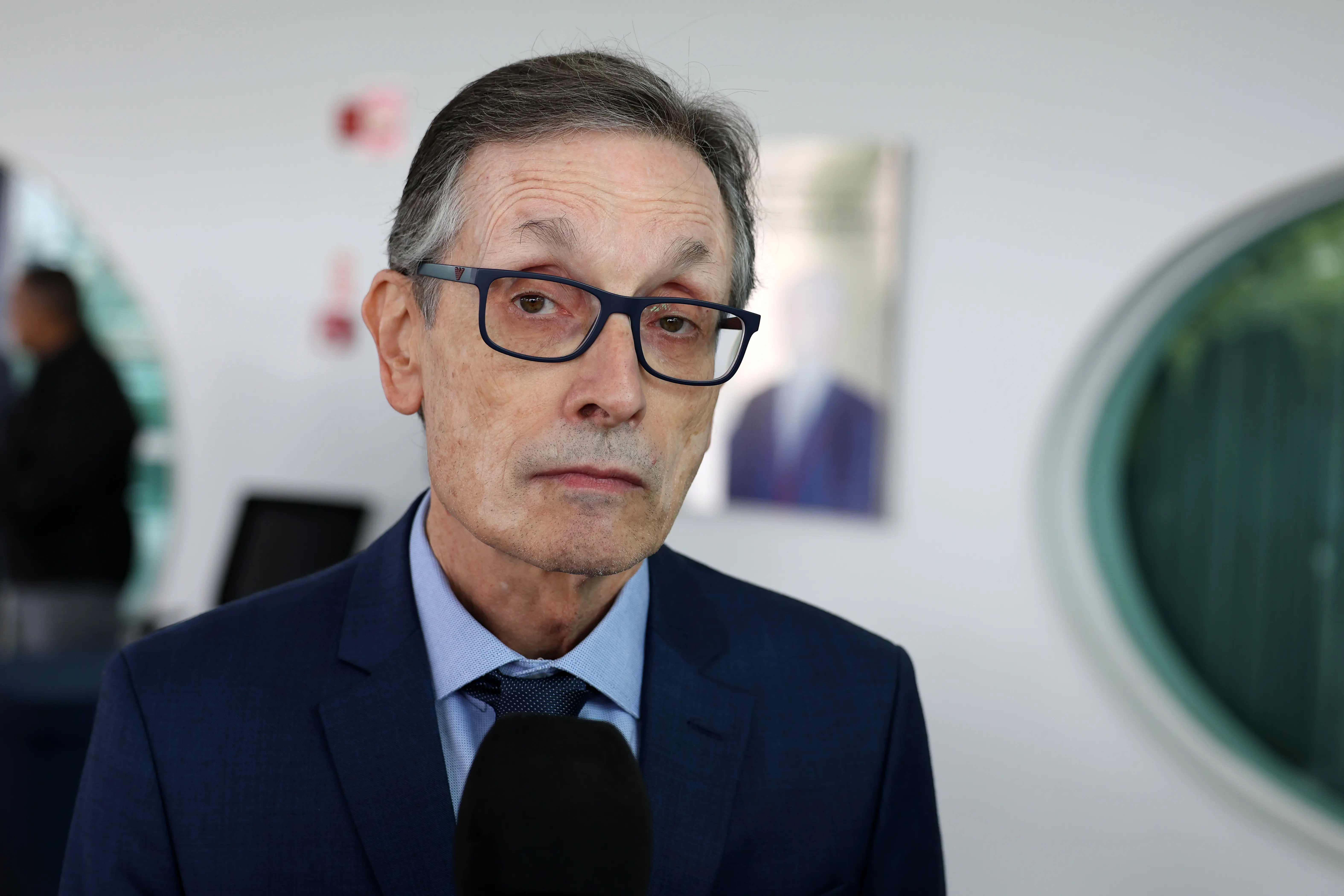Homem de pele clara, cabelos curtos grisalhos e óculos de armação preta espessa. Usa terno azul-marinho, camisa azul-clara e gravata escura. Ele olha sério em direção à câmera, com um microfone preto em primeiro plano, cobrindo o nó da gravata.