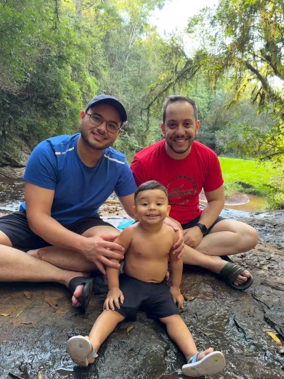 Dois homens e um menino sentados em pedras escuras na natureza. À esquerda, homem de camiseta azul e boné; à direita, homem de camiseta vermelha; à frente, o menino sorridente sem camisa.