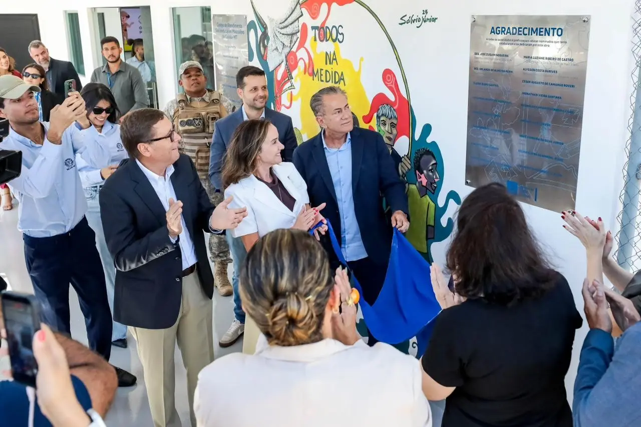 Autoridades descerrando placa de inauguração sob aplausos. Homem com terno escuro puxa pano azul de placa de acrílico. Há mural colorido e pessoas filmando e batendo palmas.