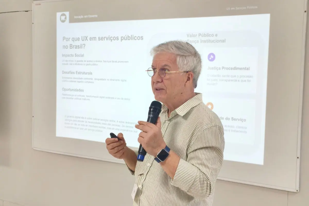Foto horizontal que mostra Álvaro Gregório, um homem de cabelos grisalhos e óculos, segurando um microfone e um controle remoto, em frente a uma tela de projeção com o texto 
