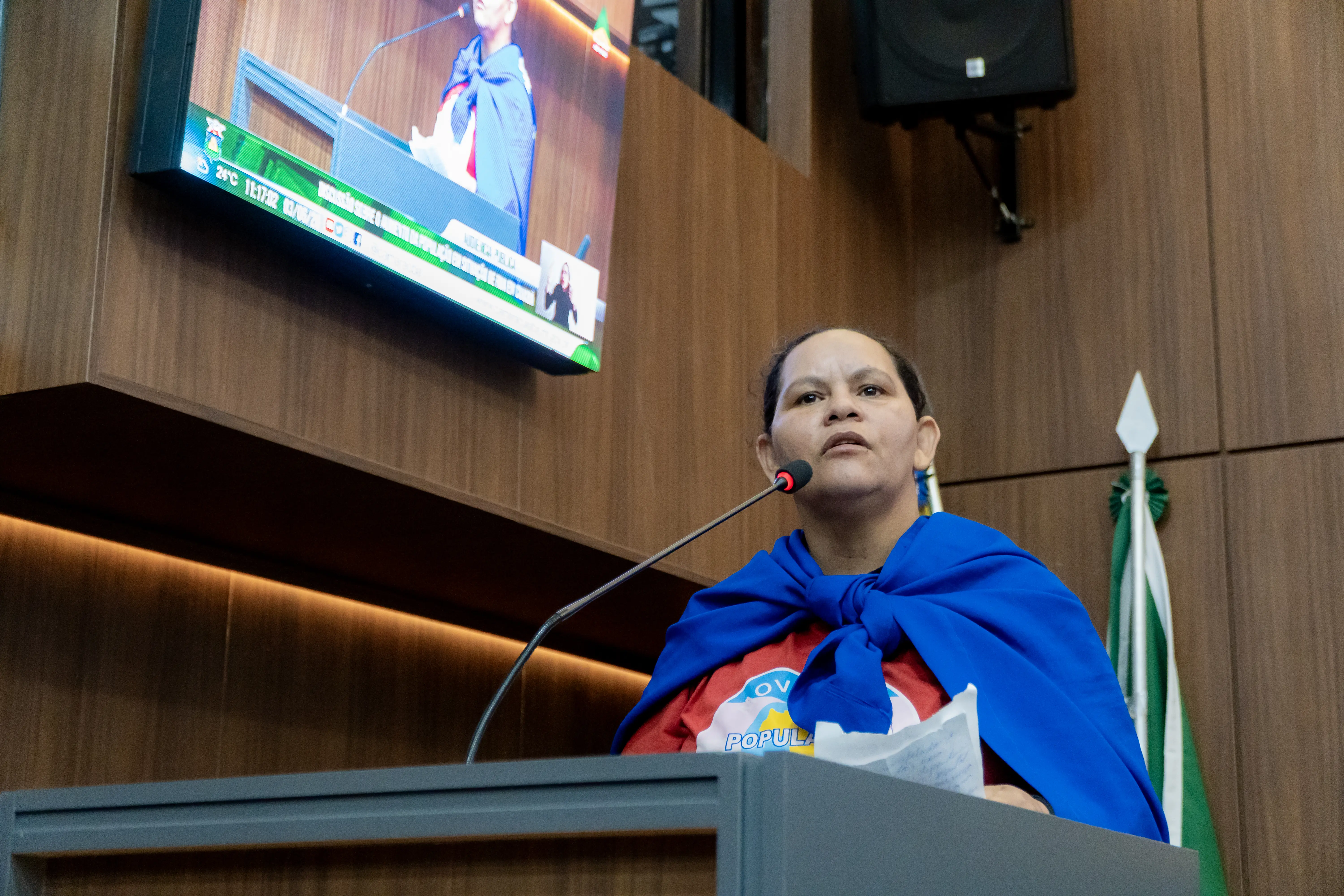 Mulher de cabelos presos fala ao microfone em um púlpito. Ela usa camiseta vermelha com estampa circular e um tecido azul amarrado sobre os ombros como capa.