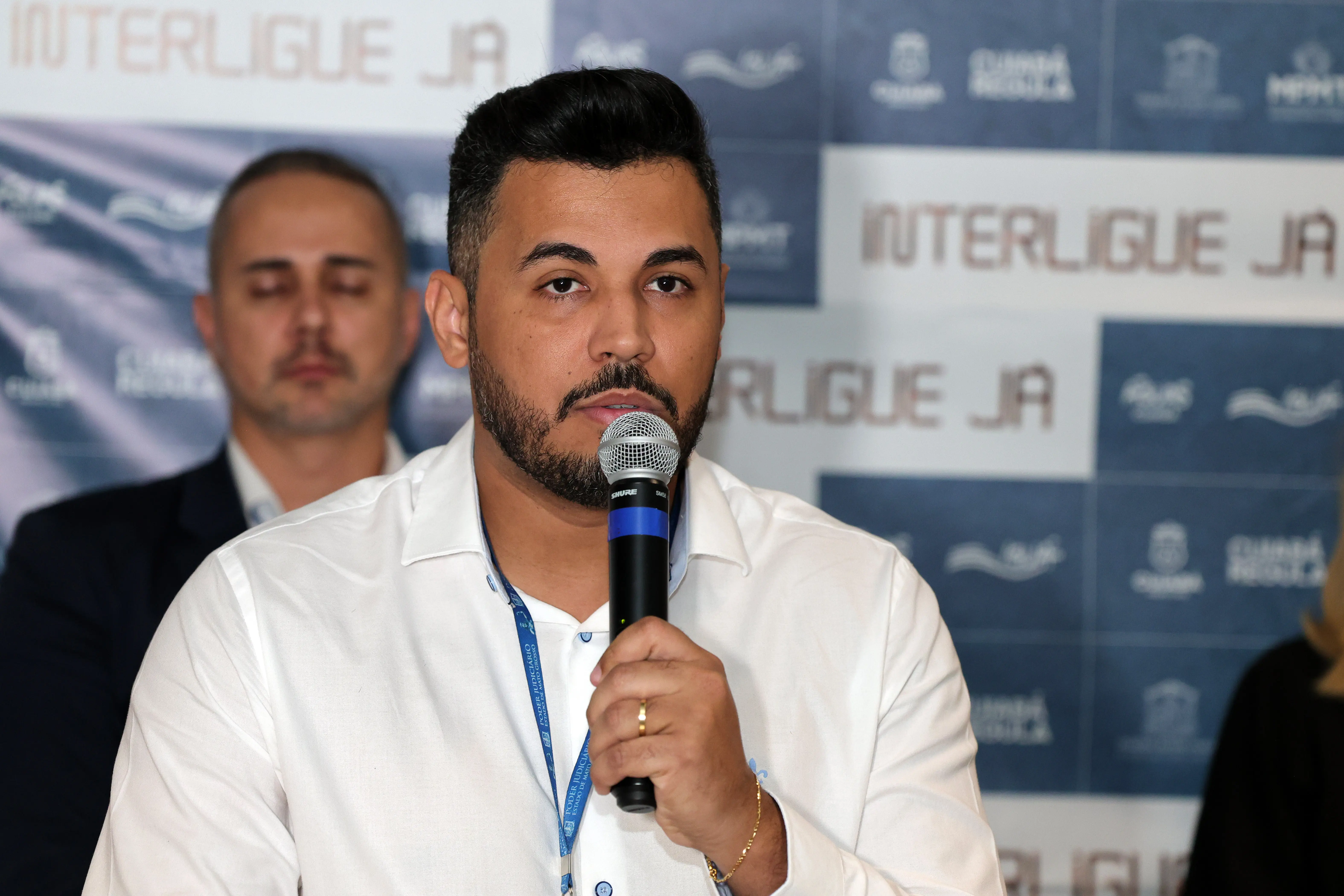 Homem de cabelos escuros e barba, veste camisa branca e crachá azul do Poder Judiciário, fala ao microfone. Ao fundo, painel do 