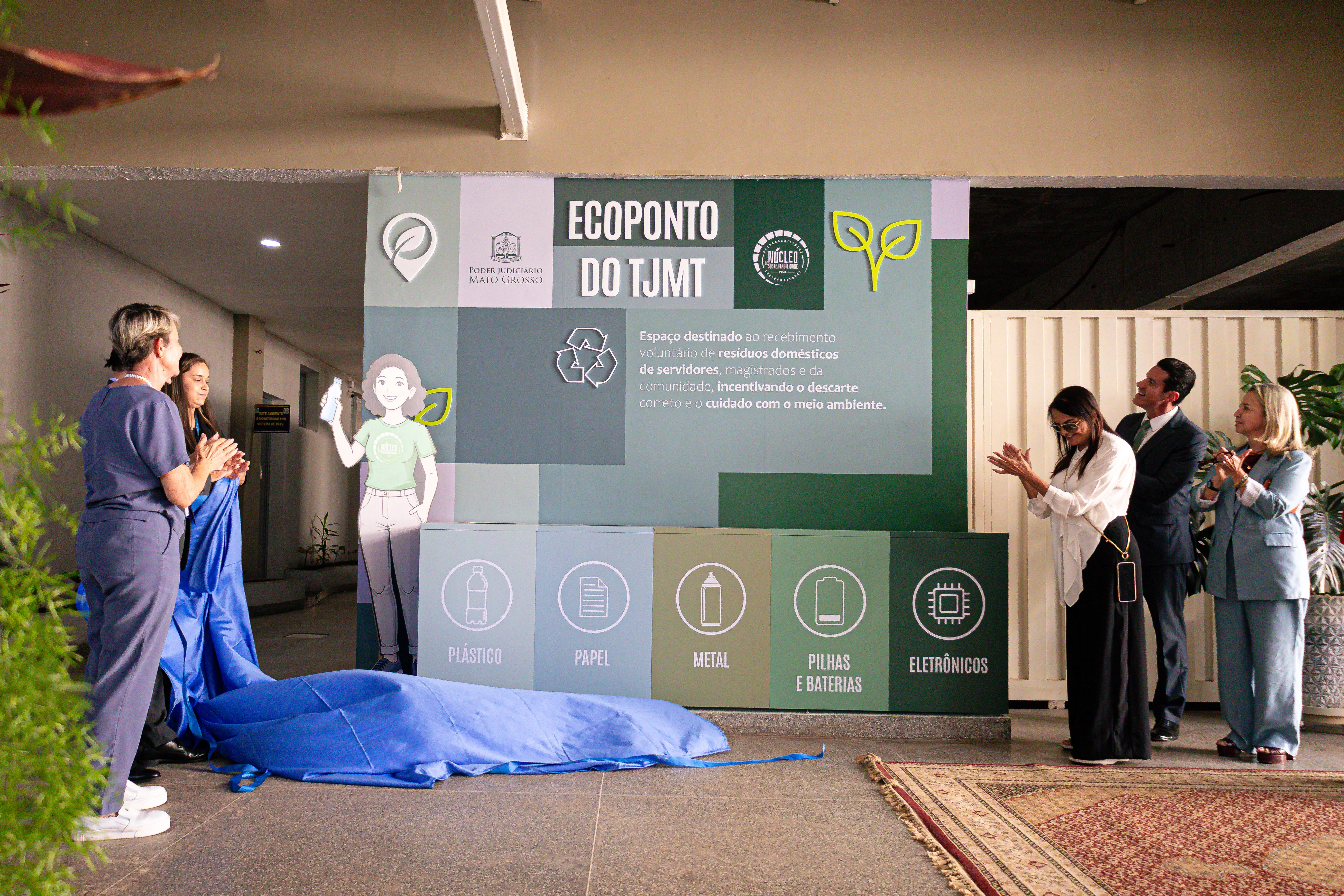 Inauguração do Ecoponto do TJMT. Pessoas aplaudem nas laterais de um grande painel verde com nichos de reciclagem para plástico, papel, metal, pilhas e eletrônicos. Um tecido azul está no chão.