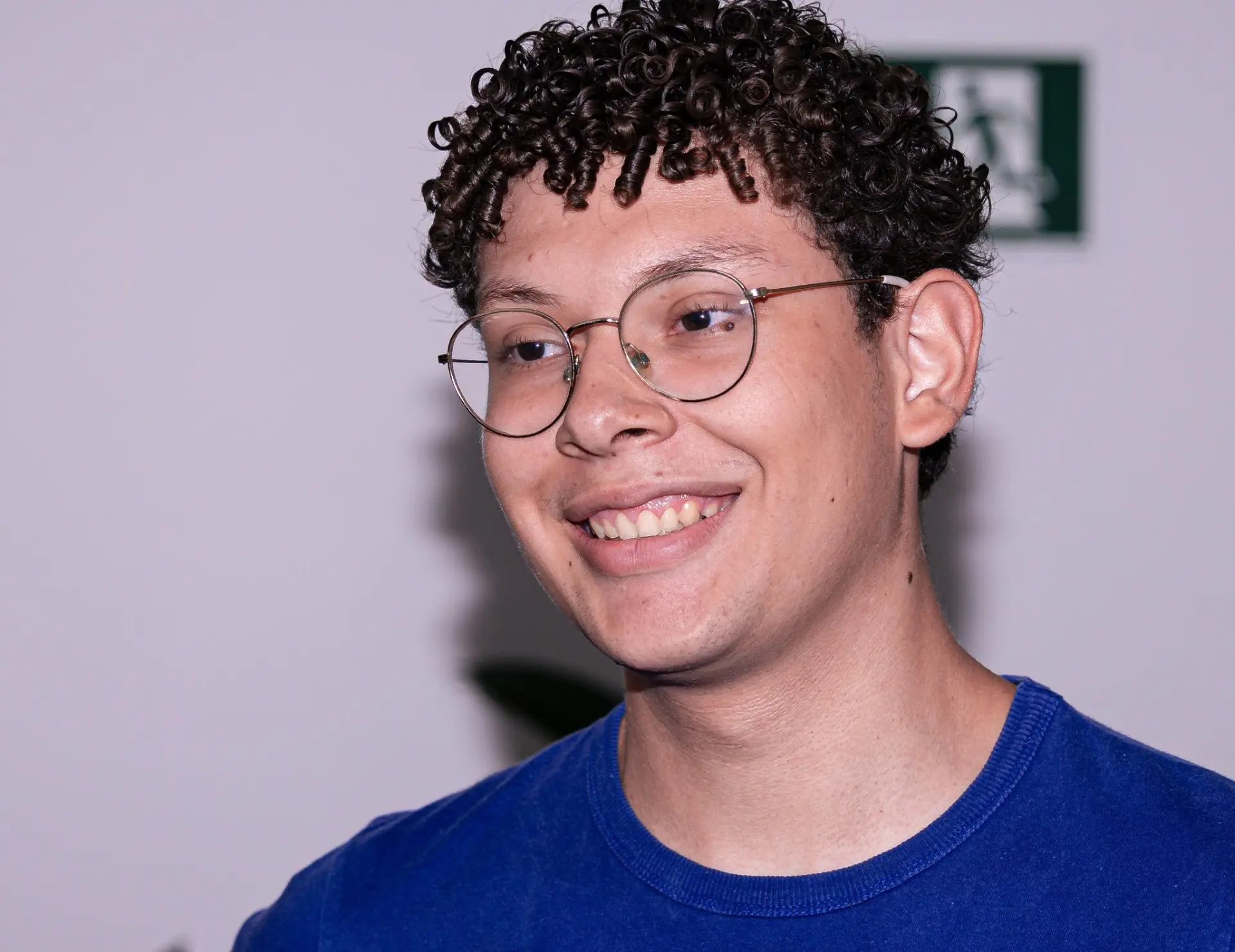 Foto horizontal em plano fechado do estagiário de Contabilidade Allan Rafael. Ele é um jovem de pele parda, olhos castanhos, cabelos castanhos, curtos e cacheados, usando óculos de grau, camiseta azul e sorrindo.