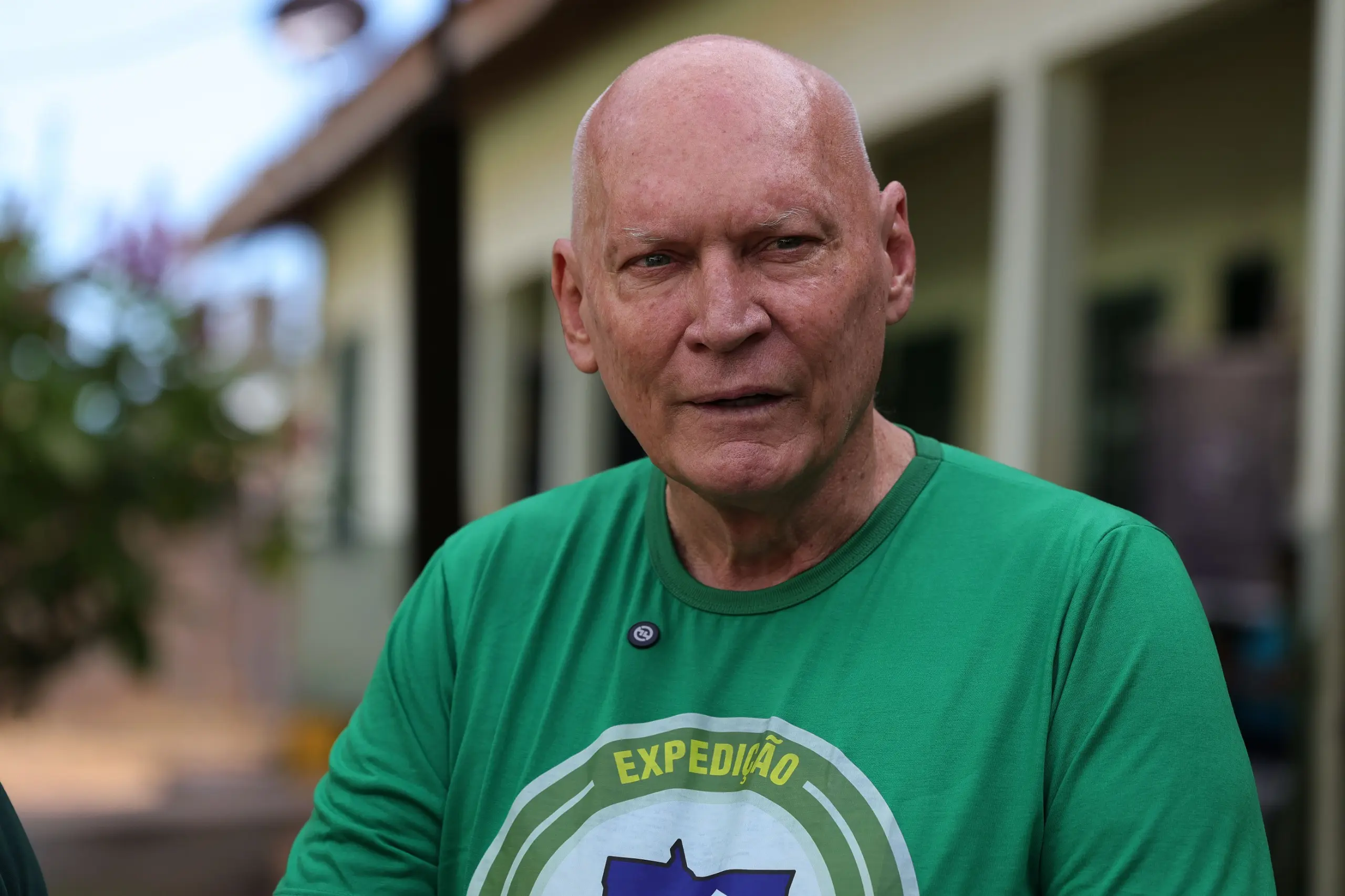 Homem idoso, de pele clara e olhos azuis, veste uma camiseta verde-clara com o texto parcial 