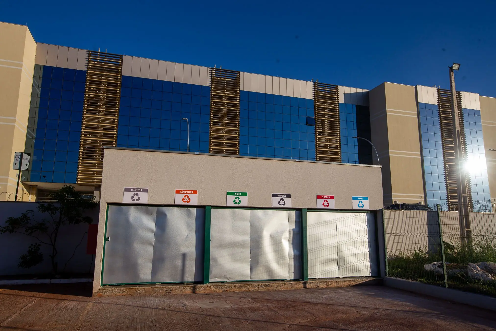 A imagem mostra a fachada do Fórum de Várzea Grande ao fundo, com estrutura moderna e vidros espelhados. Em destaque, à frente, está a central de reciclagem com compartimentos identificados para resíduos, lâmpadas, vidro, madeira, plástico e papel.