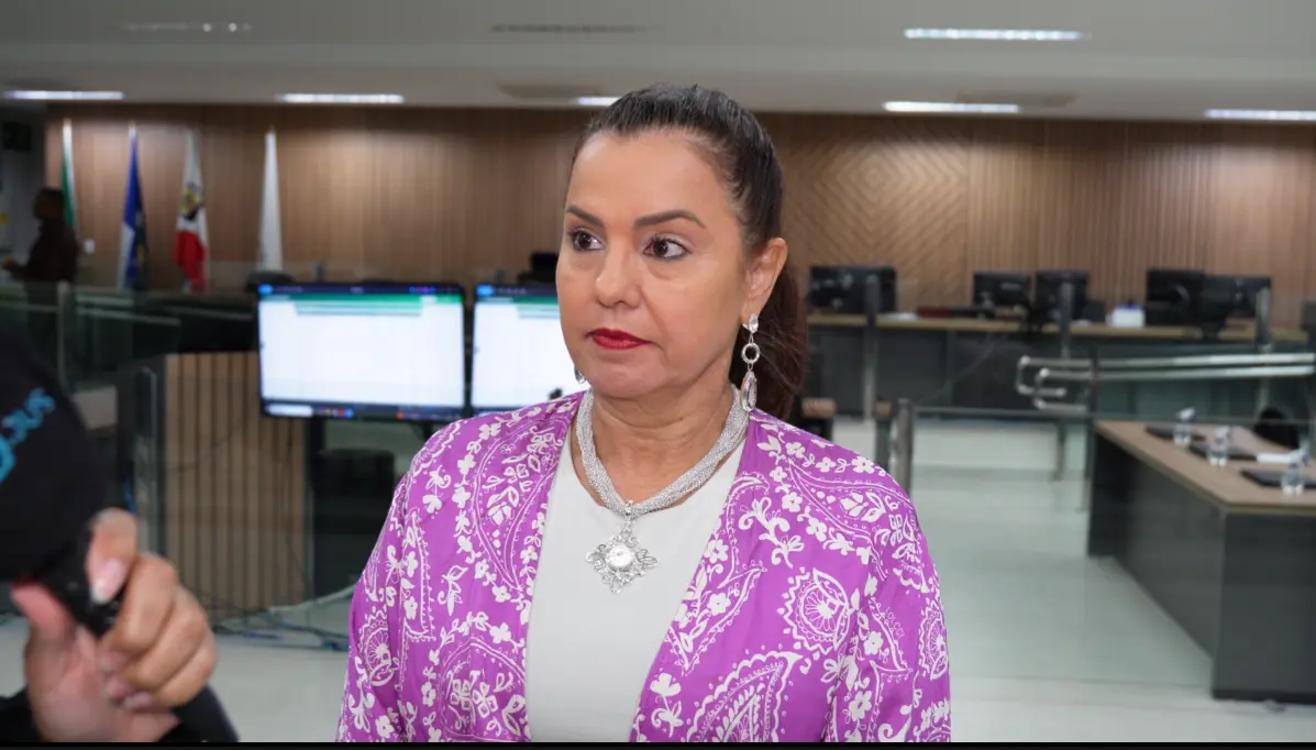 Mulher de cabelos lisos presos, veste kimono roxo floral sobre blusa branca e colar prateado, concede entrevista. Ao fundo, sala com monitores e bandeiras.